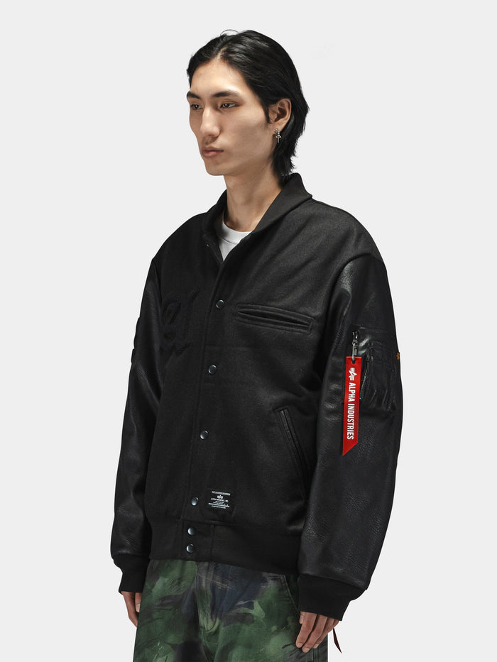 Alpha Industries Alpha Letterman Jacket in black with red Remove Before Flight tag on sleeve and green camo pants.