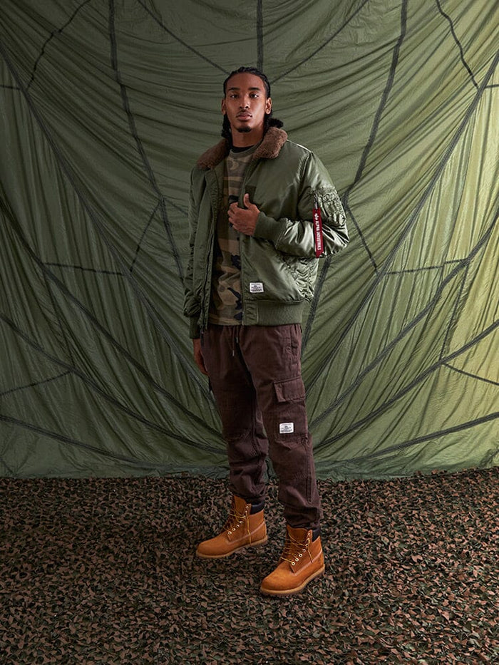Alpha Industries B-15 Mod Bomber Jacket with faux mouton collar, camo shirt, brown cargo pants, tan boots.