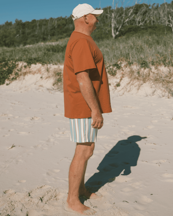 Man standing on a sandy beach wearing a brown shirt and striped shorts.