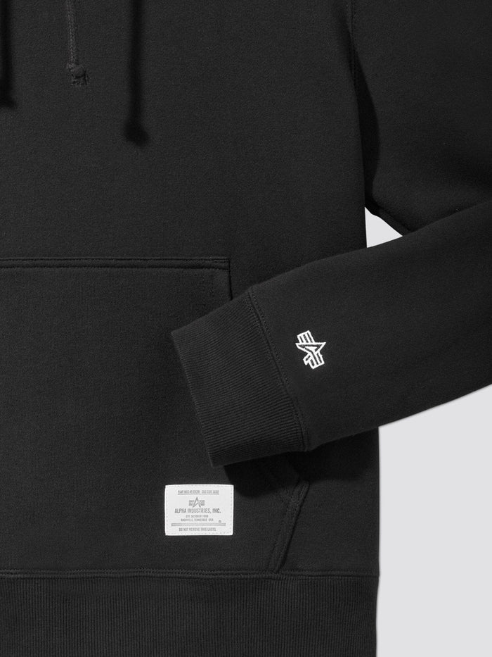 Close-up of the Black Essential Hoodie, a True Wardrobe Staple, Showcasing a Front Pocket, Logo on the Sleeve, and an Alpha Industries Label with Text Above the Pocket.