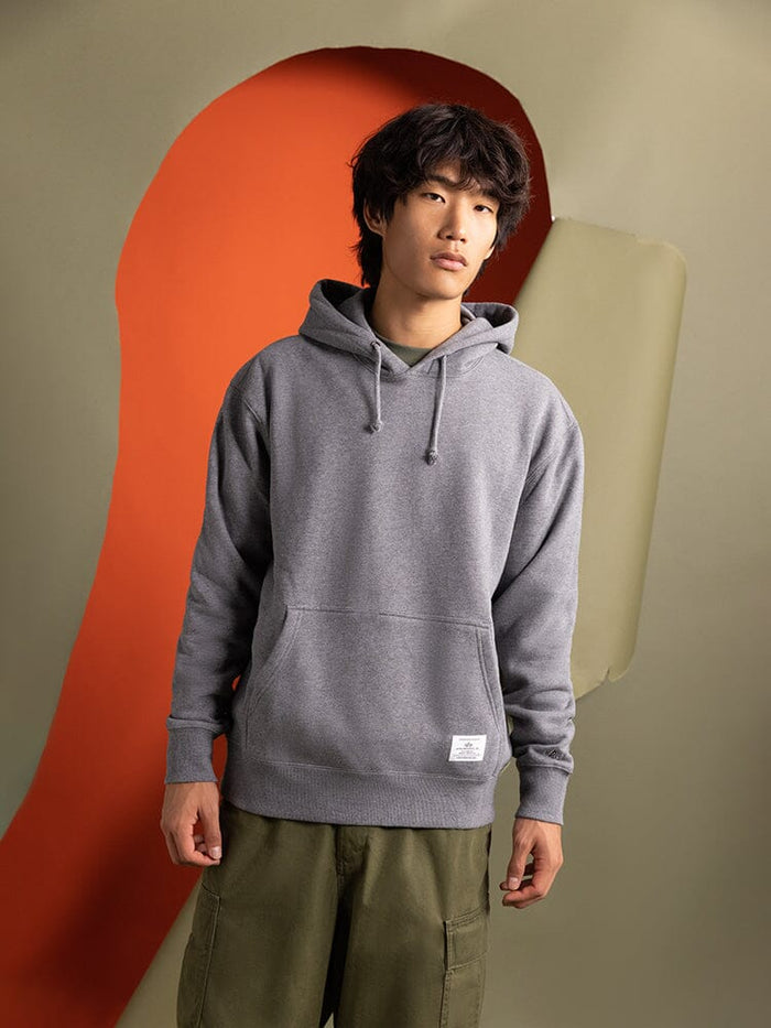 A person wearing an Essential Hoodie by Alpha Industries and green pants stands, creating a striking contrast. This unisex gray hoodie is the perfect wardrobe essential for blending style with comfort.
