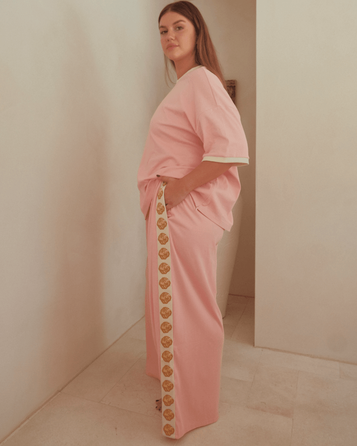 Woman wearing oversized pink shirt and wide-legged pink pants with gold shell detailing down the side. Featuring pockets in the pants.