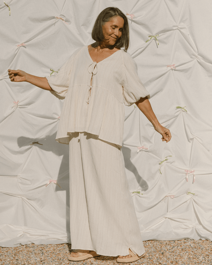 Woman in a flowing beige outfit standing against a textured white wall.