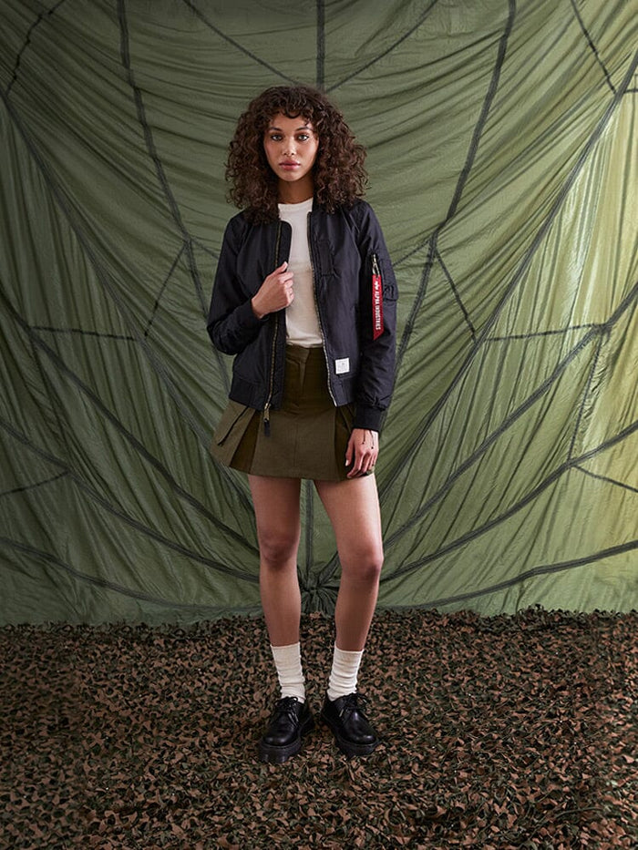 A person with curly hair stands wearing the Alpha Industries L-2B Skymaster Gen II Bomber Jacket, along with a white shirt, olive skirt, and black shoes paired with white socks.