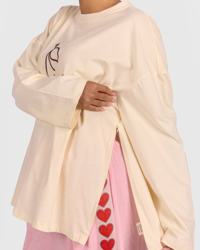 Person wearing a beige long-sleeve shirt with a black design and pink pants with red heart patterns on a white background.