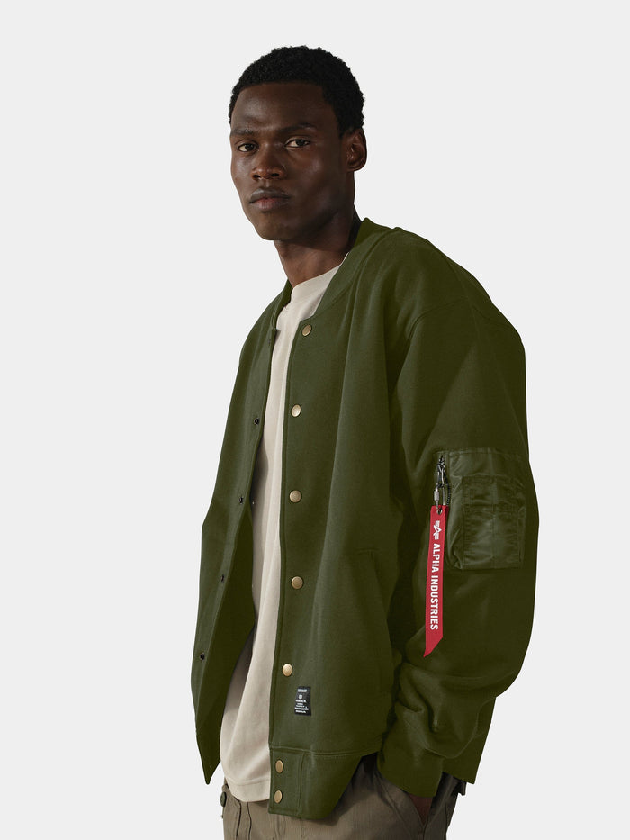Alpha Industries MA-1 Fleece Jacket in olive green with gold buttons and a red "Remove Before Flight" tag over a beige shirt.