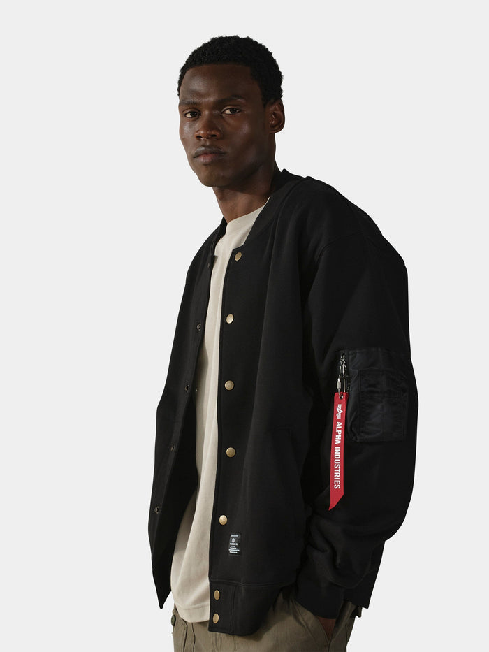 Alpha Industries MA-1 Fleece Jacket in black with a red sleeve tag, beige shirt, and khaki pants.