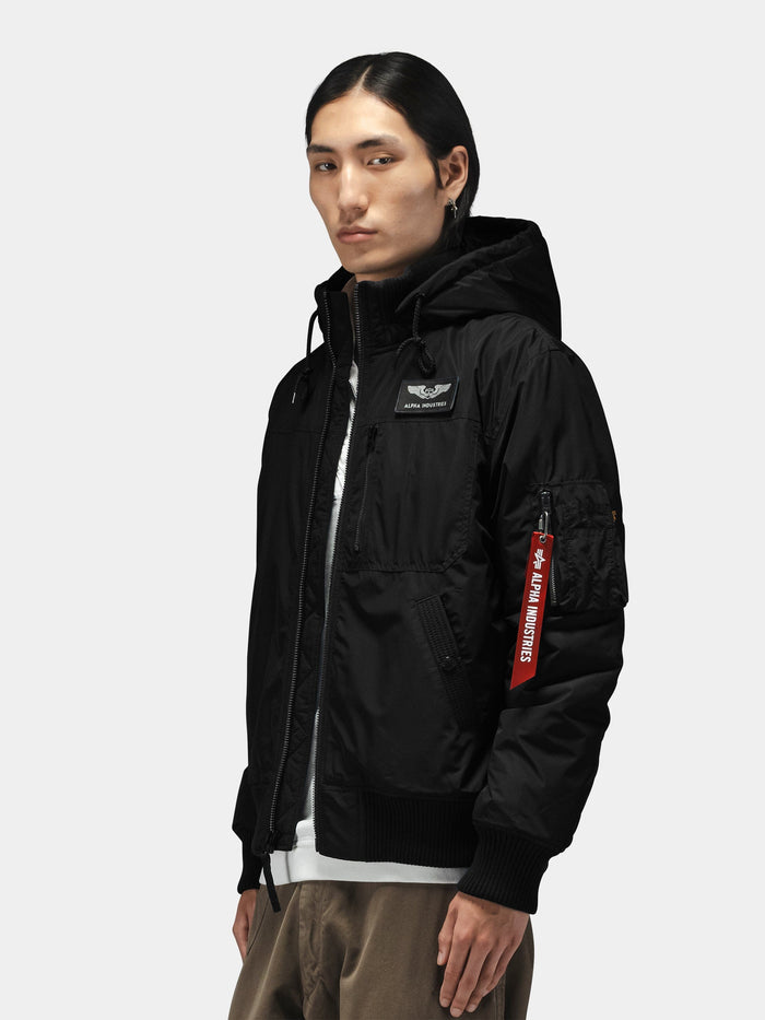 Alpha Industries Hooded Rib Flight Jacket with utility sleeve pocket, red tag, ribbed cuffs, layered over white shirt and khaki pants.
