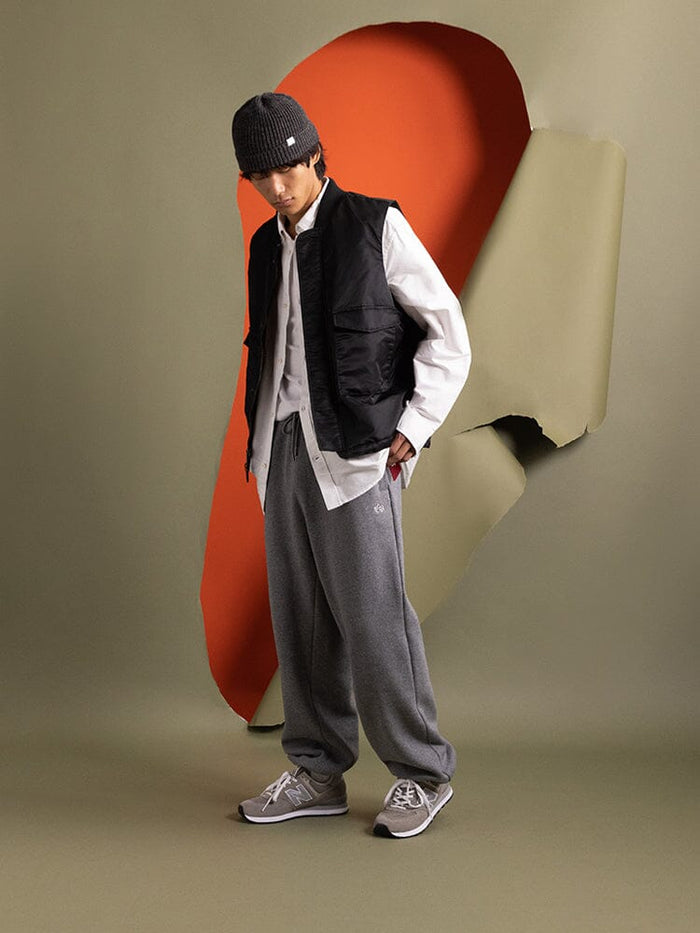 A person stands in casual attire, wearing a beanie, button-up shirt, sweatpants, and the black MA-1 Mod Vest by Alpha Industries, which features a quilted lining.