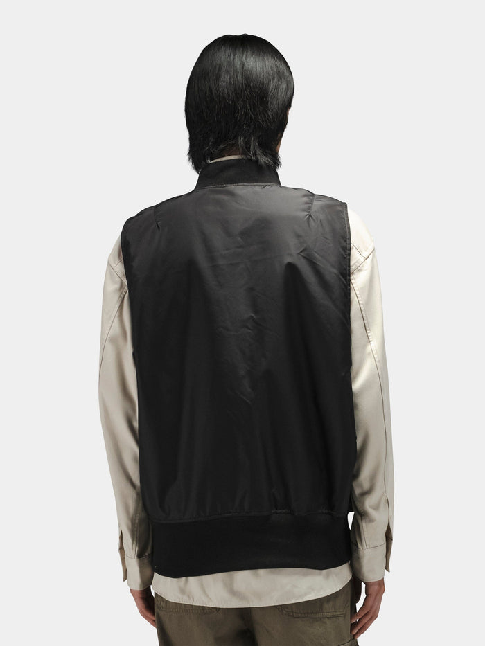 Alpha Industries MA-1 Oxford Vest in dark fabric, worn over a light long-sleeve shirt and olive pants.