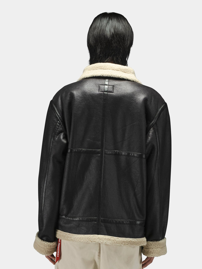 Alpha Industries Mouton B-3 black faux leather aviator jacket with beige collar and cuffs, light-colored pants.
