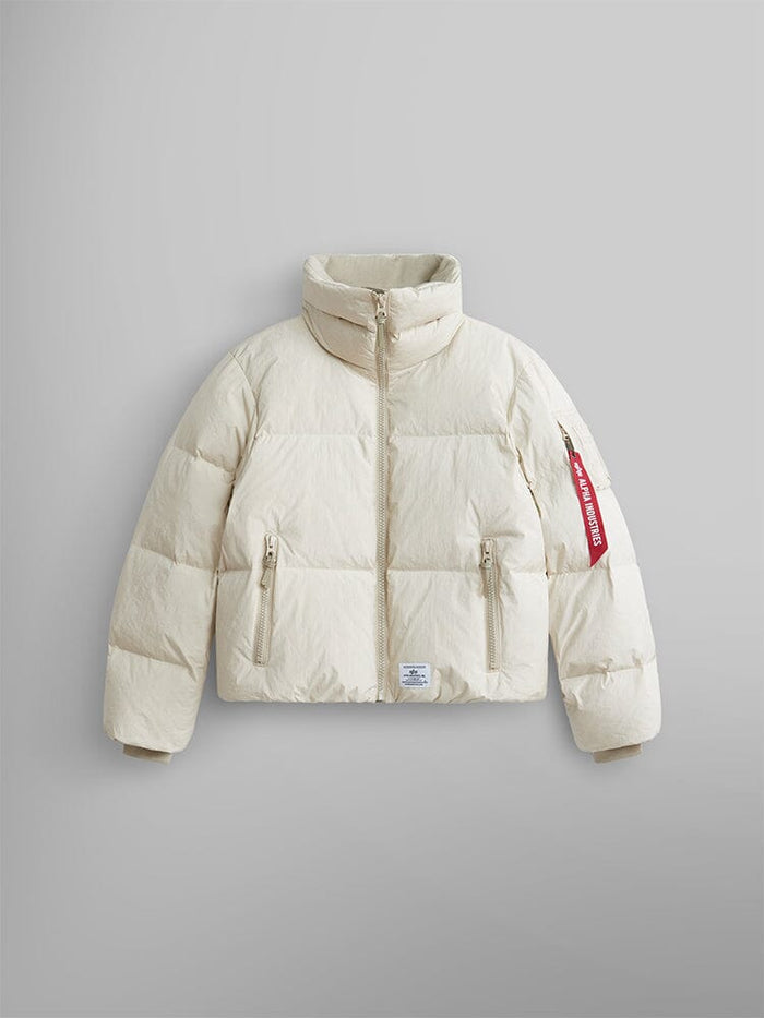 The Sierra Short Puffer W by Alpha Industries is a limestone-colored puffer parka with a high collar and water-resistant fabric. It includes two front zipper pockets and features a distinct red tag on the left sleeve.