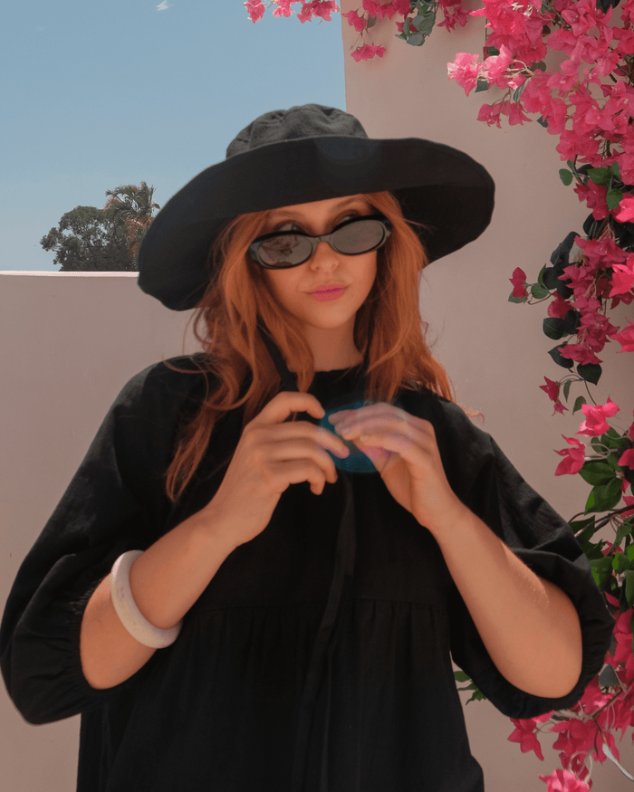 Person wearing a black hat and sunglasses, holding a colorful object, with pink flowers in the background.