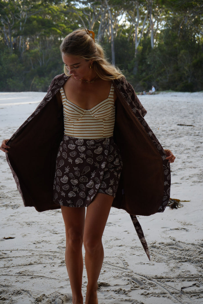 Woman standing at beach in Brown Towel Skort and Brown Seashell Towel Robe with striped swimwear on