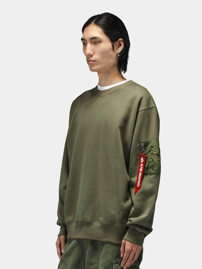 Alpha Industries Utility Pocket Crewneck in olive green with MA-1 utility pocket and red Remove Before Flight tag.