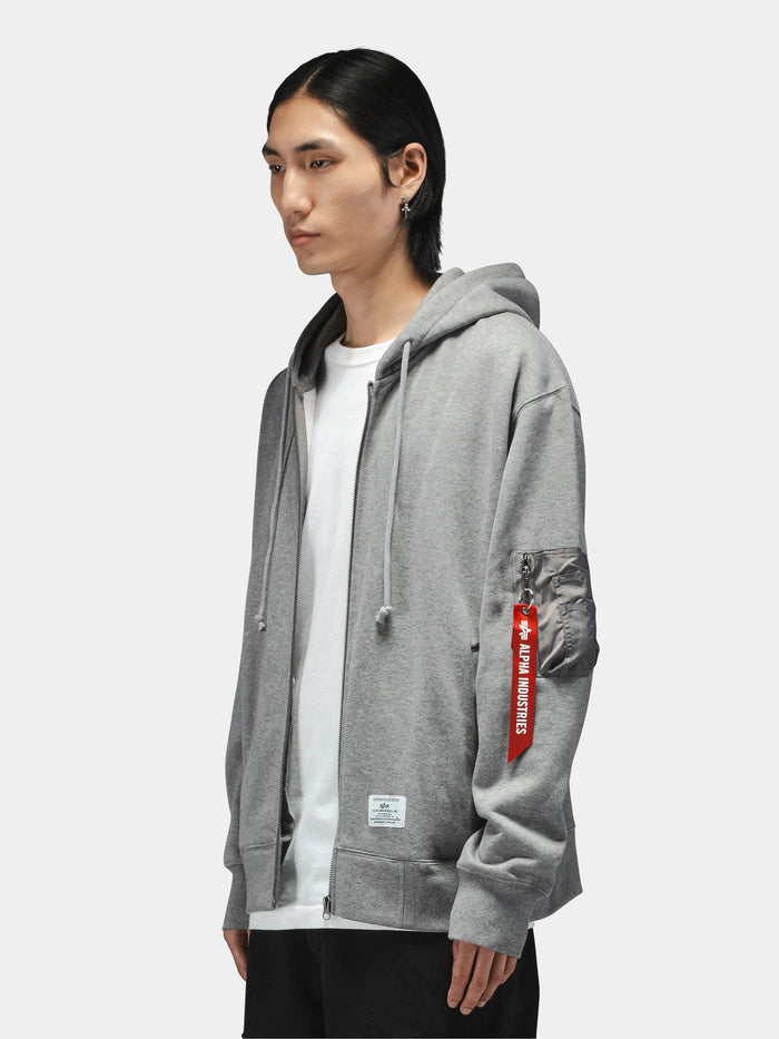 Alpha Industries Utility Pocket Full Zip Up gray hoodie with white T-shirt underneath and red Remove Before Flight tag on pocket.