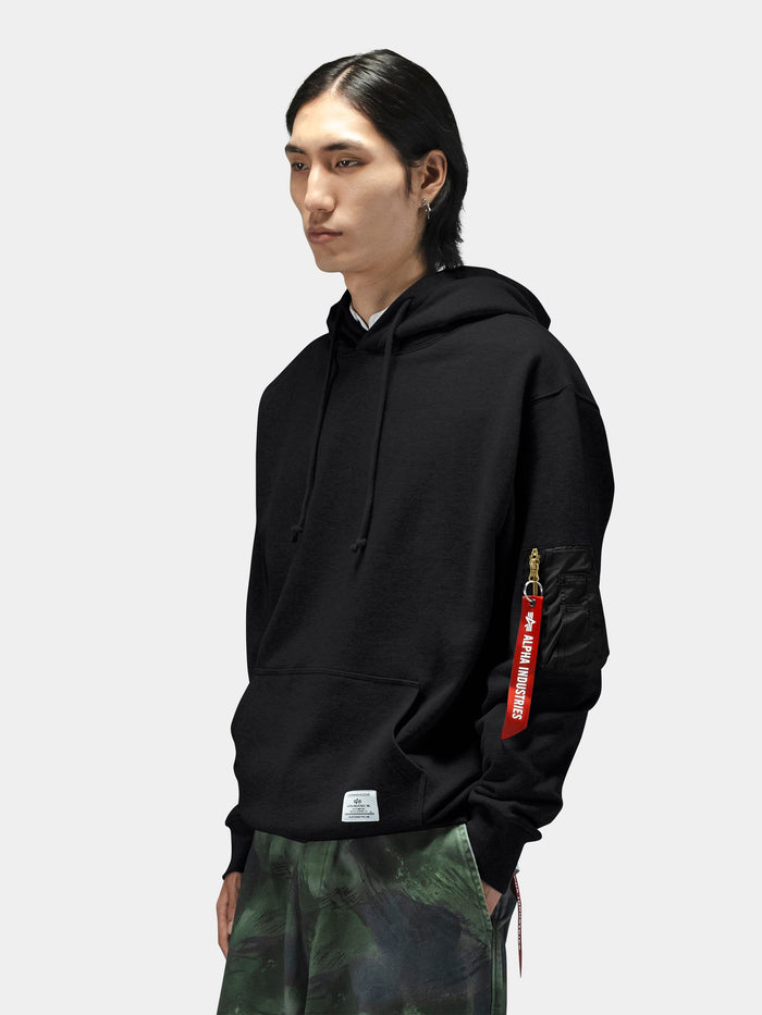 Alpha Industries Utility Pocket Hoodie in black with red Remove Before Flight tag on MA-1 sleeve pocket and green camouflage pants.